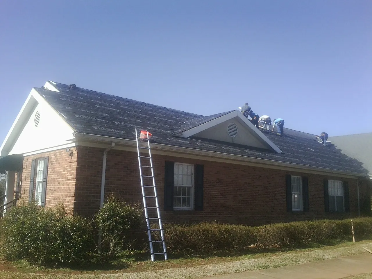 Crew tearing shingles off a brick ranch roof with a ladder set — Metal Roof Repair work by our crew in Baskerville