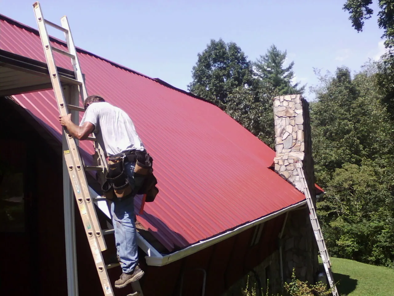 Roofer on a ladder beside a red metal roof and stone chimney — Soffit & Fascia Repair by Roofing Brothers in Baskerville